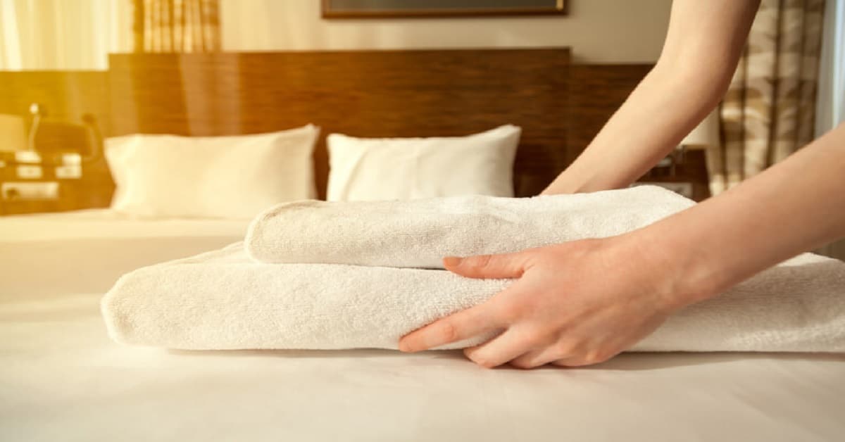 Close up of hands putting stack of fresh white bath towels on the bed sheet.