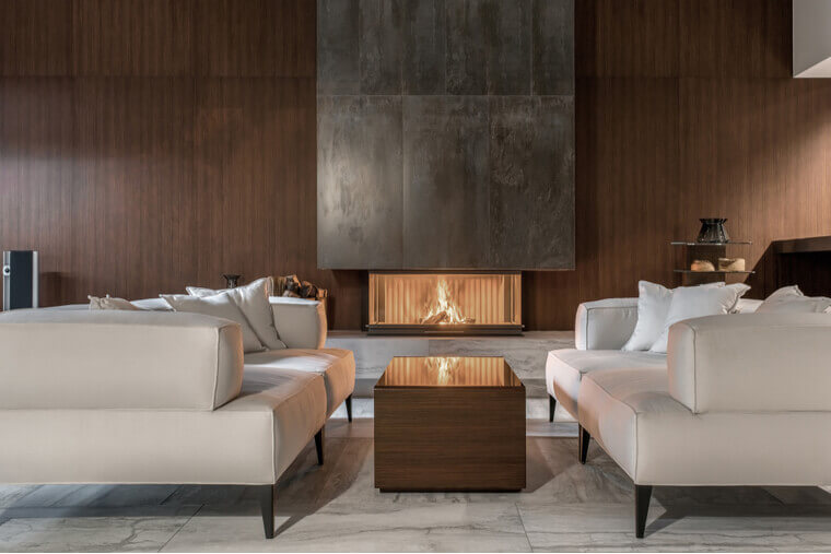 Fancy modern room with a burning fireplace, wooden wall and a tiled floor. There is a glossy table with sofas with pillows, glass stand with vases, black bag with firewood, speaker Fancy modern room with a burning fireplace, wooden wall and a tiled floor. There is a glossy table with sofas with pillows, glass stand with vases, black bag with firewood, speaker
