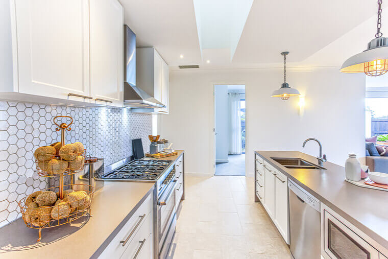 Modern kitchen with fancy items like thread balls on a small rack on the counter which has a stove next to the hallway to the door. There are hanging lamps flashing over the counter with sink and tap