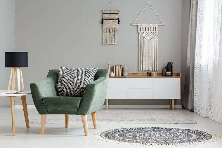 Real photo of a green armchair standing in a bright, natural living room interior with macrame hanging on gray wall above white cupboard