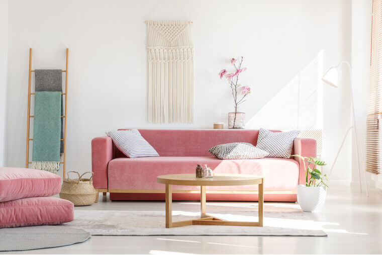 Cozy cushions and stylish textiles in a sunny, feminine living room interior with a pink, velvet sofa and a macrame on a white wall